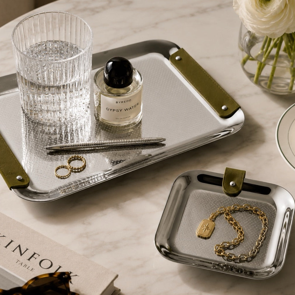 Two square silver metal jewelry and tray organizers with olive green handles, styled on a white marble table with a glass of water, perfume bottle, rings, a gold chain, and a vase of flowers.