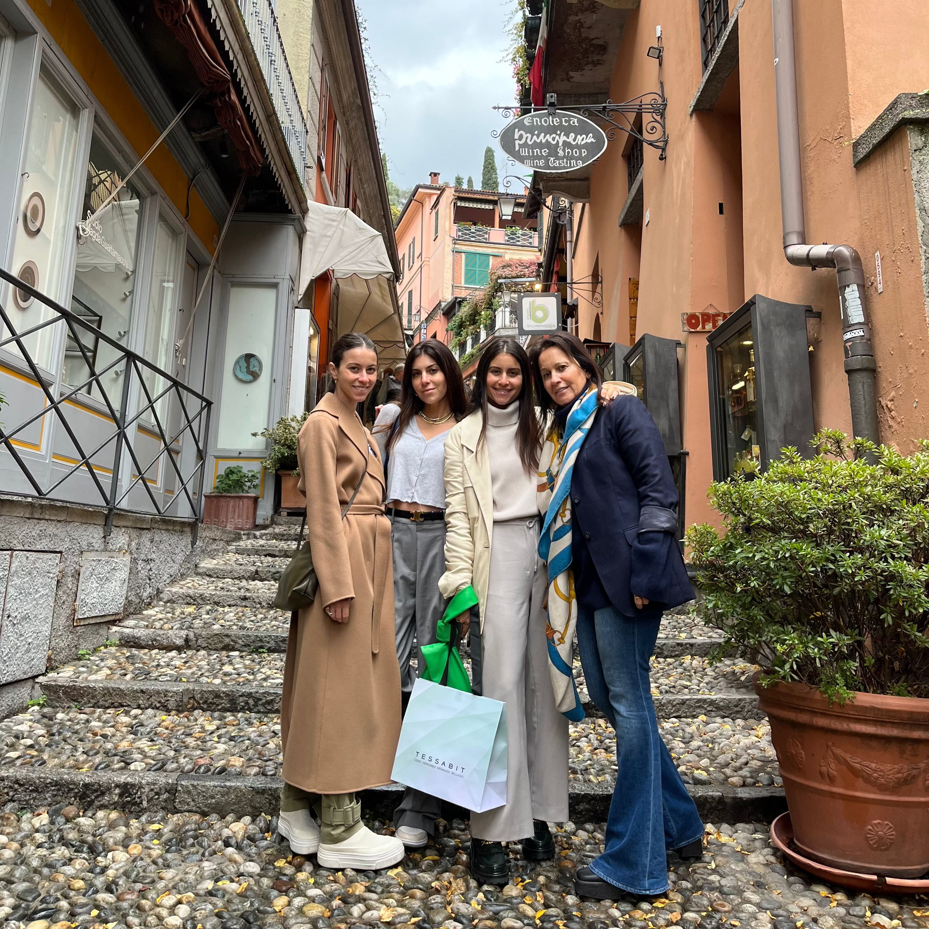 Four women posing together on stone steps in a narrow European street with orange and beige buildings, under a cloudy sky.